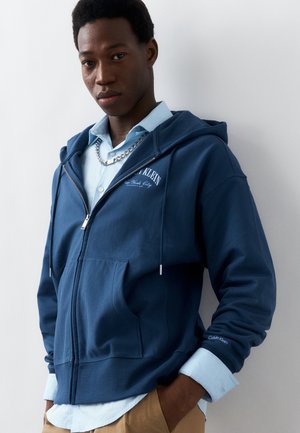 Νέος άνδρας που φοράει μπλε φούτερ με φερμουάρ Calvin Klein πάνω από ένα ανοιχτό μπλε πουκάμισο με ασημένιο αλυσίδα, στέκεται μπροστά σε έναν λευκό τοίχο.