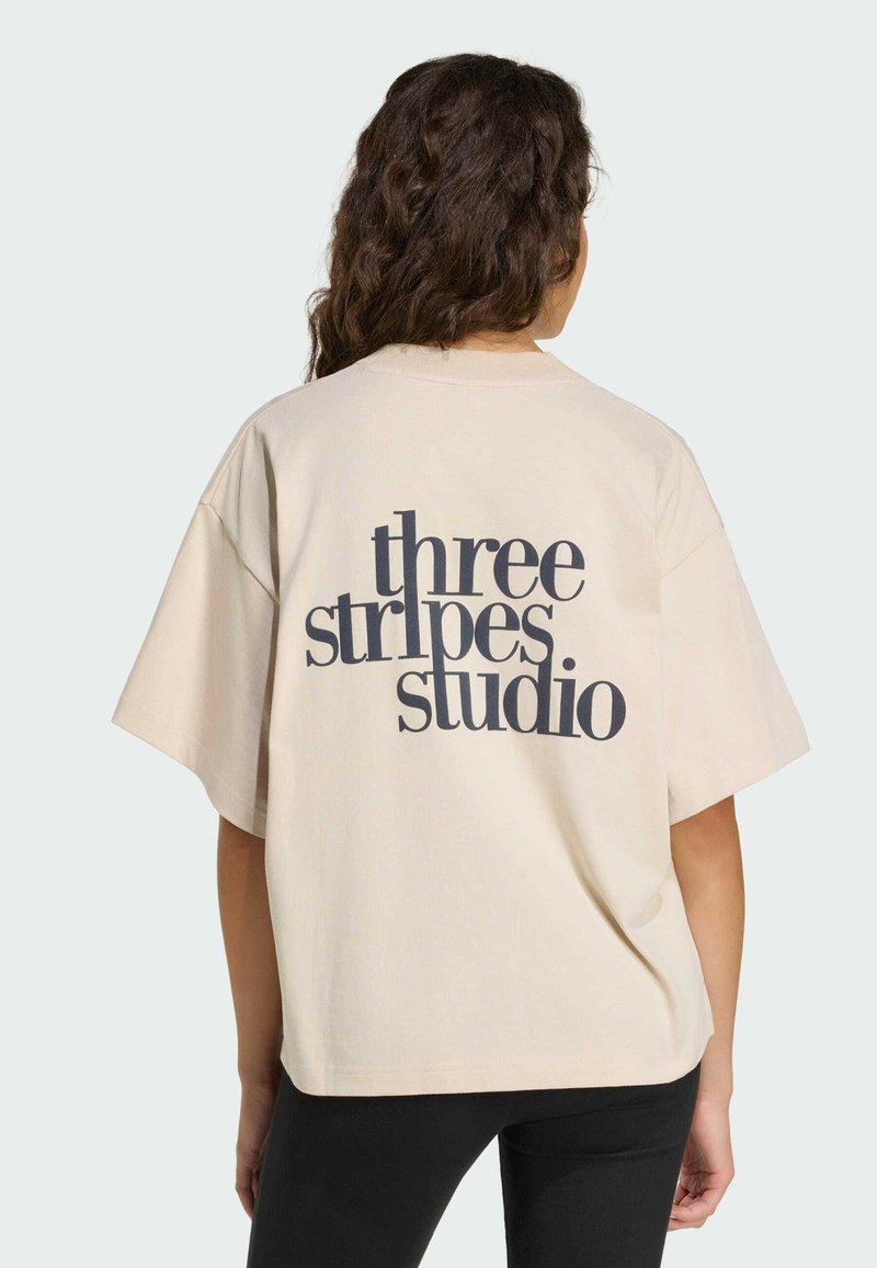 Beiges übergroßes T-Shirt mit kurzen Ärmeln, auf der Rückseite schwarzer Text "three stripes studio", glatte Baumwolltextur.