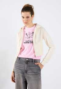 Sudadera cremosa con cremallera y textura suave sobre una camiseta rosa con un gráfico de un conejo negro. Modelo combinada con jeans de tiro alto en gris claro.