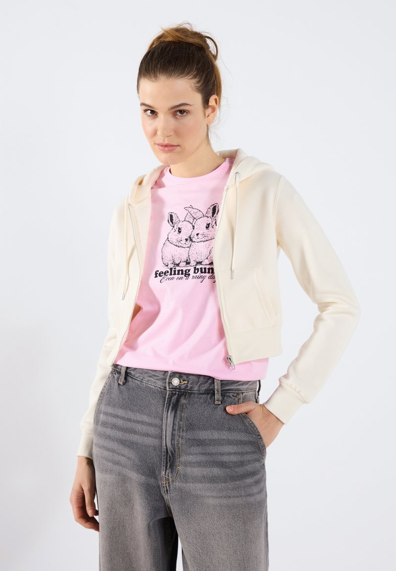 Sudadera cremosa con cremallera y textura suave sobre una camiseta rosa con un gráfico de un conejo negro. Modelo combinada con jeans de tiro alto en gris claro.