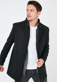 Abrigo de mezcla de lana negra con una textura suave, que cuenta con un collar, botones y un forro interior gris. El diseño es ajustado y tiene bolsillos laterales.
