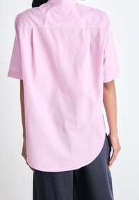 Koszula z krótkim rękawem z jasnoróżowej bawełny, z zaokrąglonym dołem, dwoma zakładkami z tyłu oraz klasycznym kołnierzykiem. Połączona z ciemnymi spodniami.