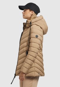 Doudoune beige avec un design en chevrons, dotée d'une capuche, de poches zippées et d'accents noirs. Portée avec une casquette noire, vue de côté.