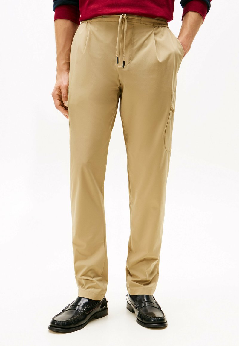 Pantalones ligeros beige con una cintura elástica y cordón, con bolsillos laterales y un corte relajado, combinados con mocasines negros.