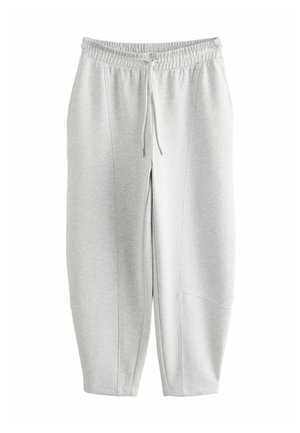 Kvinde går indendørs iført en lysegrå lynlåssweatshirt, matchende bukser med snøre og hvide sneakers mod en brun tekstureret baggrund.