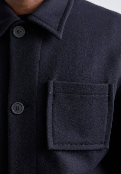 Navy wool-blend shirt jacket featuring a collar, front buttons, and a rectangular patch pocket. Soft texture with clean lines and stitching.