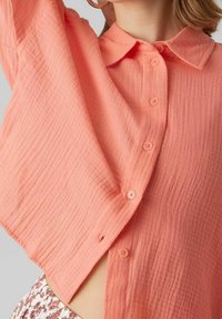 Camisa ligera de color durazno, con botones, hecha de una tela texturizada, cuello y seis botones en la parte delantera, con un corte holgado y abertura lateral.