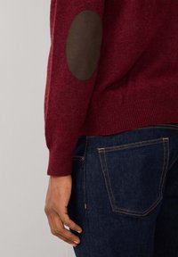 Maglione a maglia color borgogna con toppe marroni ai gomiti; polsini e orlo a coste; indossato con jeans di denim scuro con cuciture arancioni.