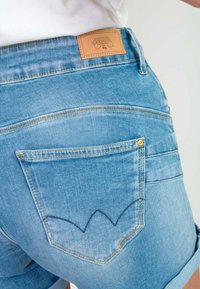 Shorts de mezclilla en azul claro con un acabado texturizado. Presenta una etiqueta en la cintura y un bolsillo trasero con detalles de costura curva.