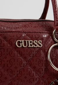 Gros plan sur un sac à main matelassé bordeaux brillant avec motif embossé, logo argenté "GUESS" et anneaux métalliques sur la poignée.