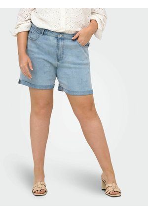 Žena v svetlomodrých denimových kraťasoch s hviezdičkovým vzorom, bielom blúzke s dierkovaným vzorom a béžových sandáloch na opätku, stojaca s jednou rukou v vrecku.