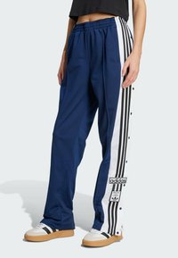 Navy blue Adidas track pants with white side panels, black stripes, snap buttons, and brand logos worn with white and black sneakers.