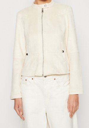 Persona che indossa una giacca color panna con zip, colletto a scatto e pannelli testurizzati, abbinata a jeans bianchi, in piedi su uno sfondo semplice.