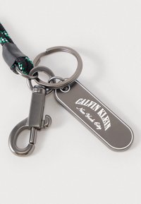 Μαύρο και πράσινο πλεκτό κορδόνι με μεταλλικό κρίκο για κλειδιά και κλιπ. Συνημμένη μεταλλική ετικέτα με εγχάρακτο το κείμενο «CALVIN KLEIN» σε λευκό χρώμα. Ομαλή υφή.