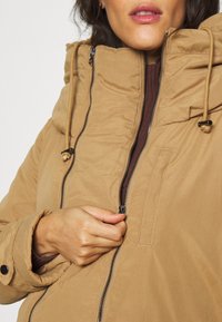 Femme portant une doudoune beige à col montant, tenant la fermeture éclair tout en la fermant partiellement.