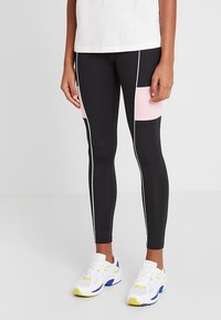 Zwarte leggings met roze panelen, witte zijkanten, slim fit en rekbare stof. gedragen met witte en gele sportschoenen.