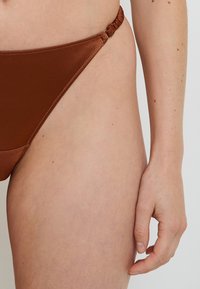 Bas de bikini marron en tissu lisse, avec un détail froncé sur le côté et une taille basse, idéal pour la plage.