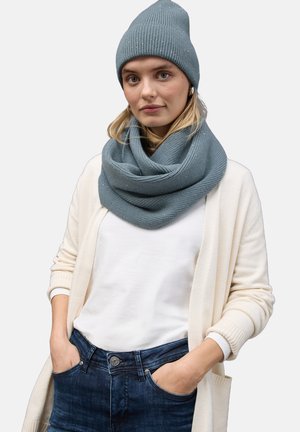 Bonnet en tricot gris et écharpe infinie assortie, tous deux présentant une texture douce et un design côtelé, associés à un haut blanc et un cardigan crème.