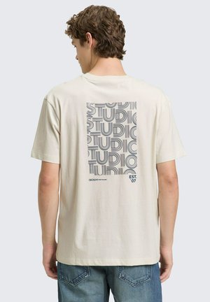 Junger Mann mit lockigem Haar trägt ein elfenbeinfarbenes T-Shirt mit wiederholtem "STUDIO"-Schriftzug und "DENIM Tom Tailor EST. '07" auf der Rückseite.