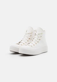 High-top white sneakers made of canvas with gold eyelets, rubber sole, and textured toe cap, featuring flat white laces and a seamless design.