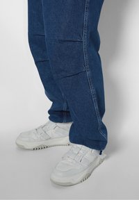 Vita sneakers med en texturerad ovandel och justerbara remmar, tillsammans med mörkblå jeans med synliga sömmar och avslappnad passform.