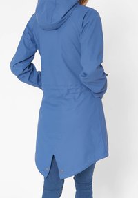 Blauwe waterdichte jas met een capuchon, verstelbare taille en een langere achterkant. Gladde textuur, minimale hardware en een driehoekige accentrand aan de onderzoom.