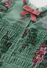 Tessuto floreale verde arricciato con piccoli bottoni verdi e un fiocco in velluto rosa vicino a un colletto trasparente.