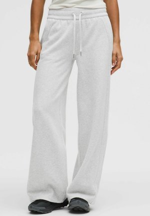 Frau trägt hellgraue, weit geschnittene Jogginghosen mit Kordelzug in der Taille und schwarze Sportschuhe, steht vor einem schlichten Hintergrund.