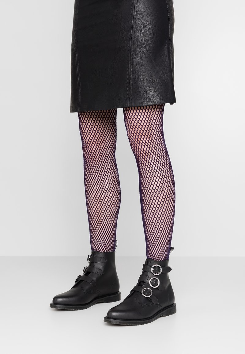 Jambes portant des collants résille noirs, une jupe en cuir noire et des bottines noires avec trois boucles argentées sur chaque botte.