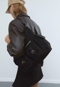 Femme portant une veste en cuir marron et une casquette beige, portant un grand sac noir avec des poches à boucle sur son épaule.