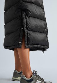 Melna pūkainā jaka ar ieaustingām frontēm, spiedpogu aizdari un garumu līdz puslaim. Apvienota ar pelēkiem sporta apaviem ar logotipu un sieta akcentiem.