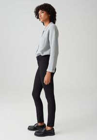 Vrouw met krullend haar, gekleed in een lichtgrijs geribd jasje, zwarte strakke broek en zwarte veterschoenen, staande opzij tegen een neutrale achtergrond.