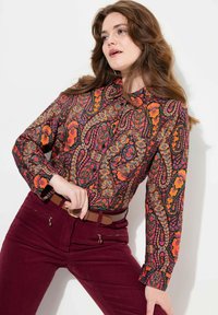 Long-sleeve patterned blouse featuring floral designs in vibrant orange, pink, and green on a black background. Paired with maroon pants.