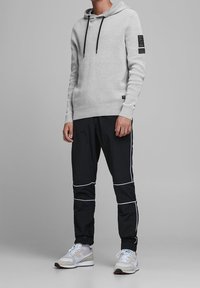Sudadera con capucha de punto gris claro con cordón y parche negro; combinada con pantalones deportivos negros con rayas blancas y zapatillas claras.