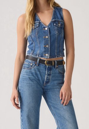 Woman wearing buttoned sleeveless denim vest and high-waisted blue jeans with black belt, standing against plain background.