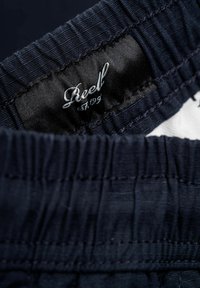 Cintura elástica azul marino con tejido texturizado, con una etiqueta tejida que muestra "Reel est. 1999" en bordado plateado.