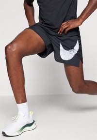 Mustat urheilushortsit, joissa on suuri valkoinen swoosh-logo, valmistettu kevyestä materiaalista. Yhdistetty valkoisten sukkien ja valkois-vihreiden tennarien kanssa.
