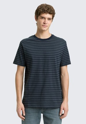 Junger Mann, der ein dunkelblaues und schwarzes, horizontal gestreiftes T-Shirt mit kurzen Ärmeln und hellblaue Jeans trägt, vor einem einfarbigen Hintergrund steht.