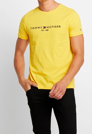 Żółta bawełniana koszulka z krótkim rękawem i okrągłym dekoltem, z logo "TOMMY HILFIGER" w kolorze czarnym i czerwonym. Noszona z czarnymi spodniami.
