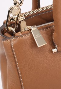 Bruine leren handtas met gouden hardware, bovenrits en gedetailleerde stiksels. Bevat een metalen label met "Paul Costelloe" gegraveerd.