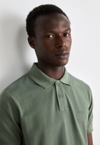 Grønn polo-skjorte laget av bomull, med krage, to-knapp placket, og subtile broderte logo på brystet. Glatt tekstur.