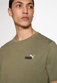 Μπλούζα από βαμβάκι σε ελιά χρώμα με στρογγυλή λαιμόκοψη, διακοσμημένη με κεντητό λευκό λογότυπο Puma και μαύρο κείμενο στο στήθος.