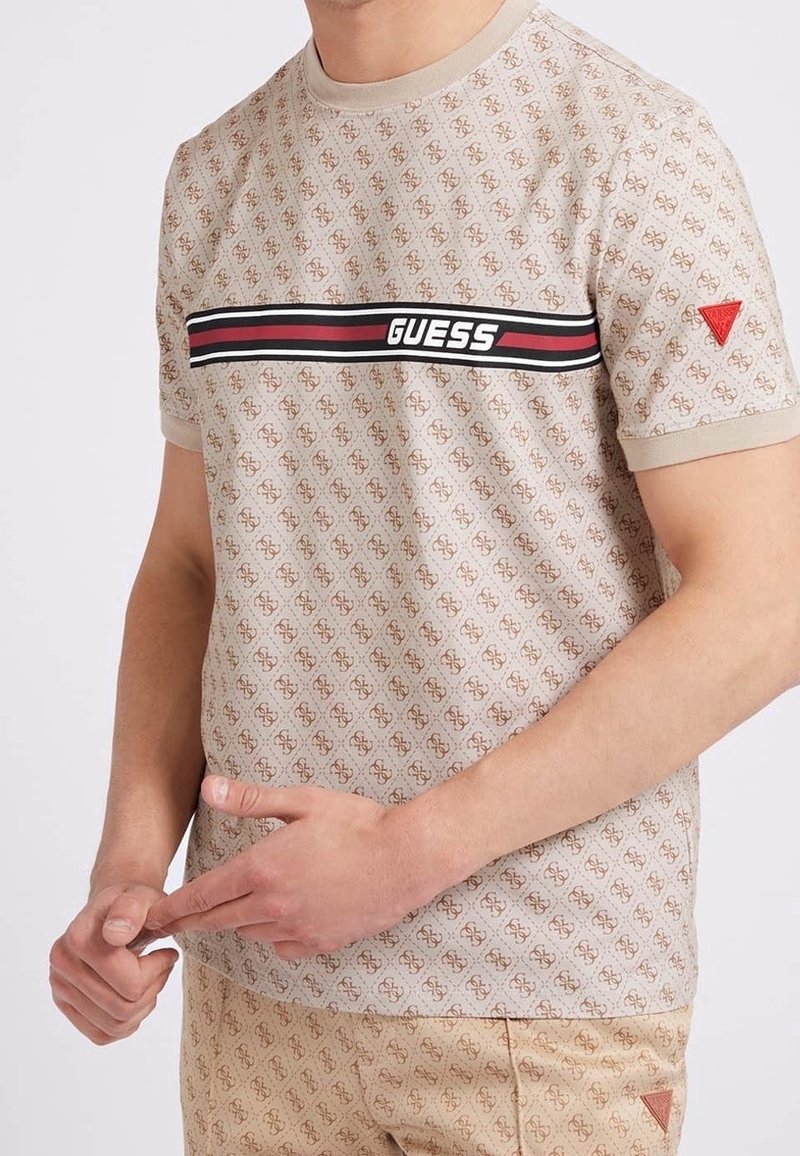 Homme portant un t-shirt beige Guess avec un motif logo intégral et un short assorti, mains jointes sur la taille.