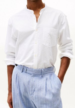 Mężczyzna noszący białą, teksturowaną koszulę zapinaną na guziki z podwiniętymi rękawami oraz jasnoniebieskie plisowane spodnie z szlufką na pasek.