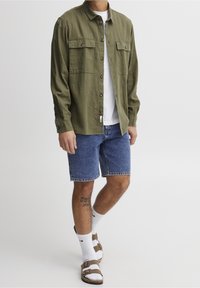 Camicia a maniche lunghe verde oliva con due tasche sul petto, abbinata a pantaloni corti in denim blu, t-shirt bianca, calzini bianchi e sandali marroni.