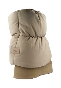 Bota acolchada beige con una parte superior esponjosa, textura suave y suela de goma. Presenta un parche con logo en el lateral y un tacón ligeramente elevado.