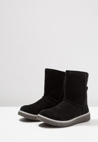 Pár černých semišových bot do půli lýtek s plochými šedými gumovými podrážkami na bílém povrchu na pozadí jednoduché světlé šedé barvy.