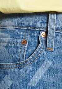 Světle modré džínové kalhoty s viditelnými kapsami a měděnými nýty. Mají oranžové švy a strukturovaný povrch s vybledlým vzorem.