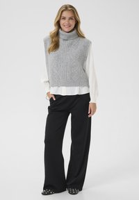 Sivi pleteni bezrukavnik s turtleneck izrezom, ribanom teksturom i dijamantnim uzorkom, uparen s bijelom košuljom dugih rukava i crnim širokim hlačama.