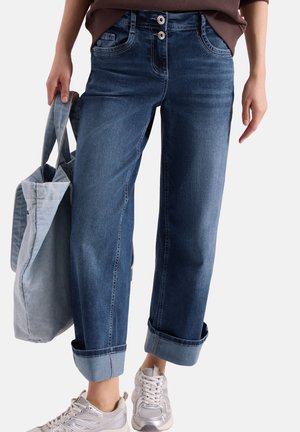 Personne portant un jean bleu foncé retroussé et des baskets blanches, tenant un grand sac cabas en denim bleu clair d'une main.